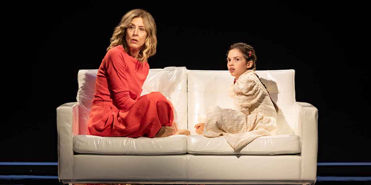 A person in a red dress and a child in a white outfit sit on a white sofa, looking concerned.
