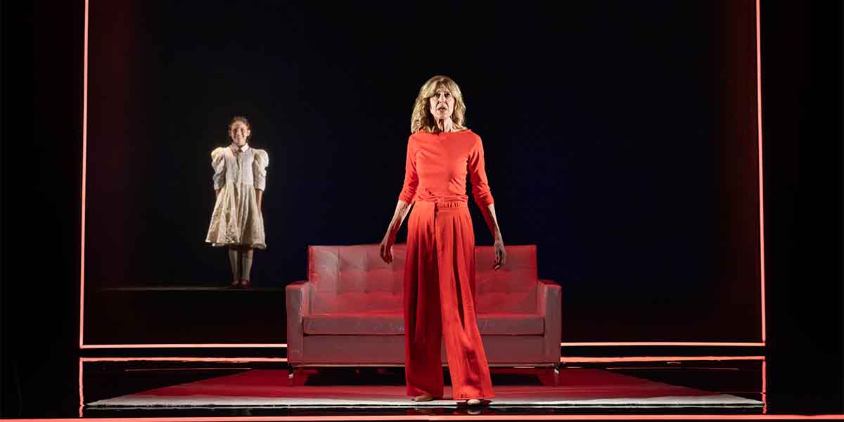 A person in a red outfit stands confidently in front of a sofa, with a child in white behind her.