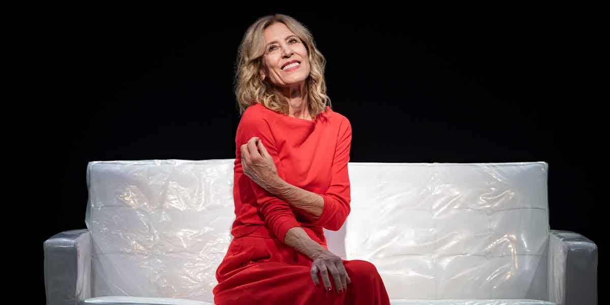 A person in a red dress smiles while sitting on a white sofa against a dark background.