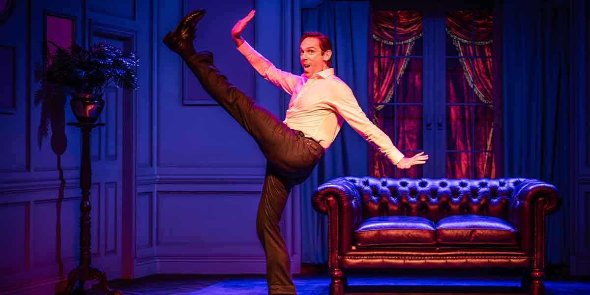 A man in a white shirt and black trousers performs a high kick beside a brown leather sofa.