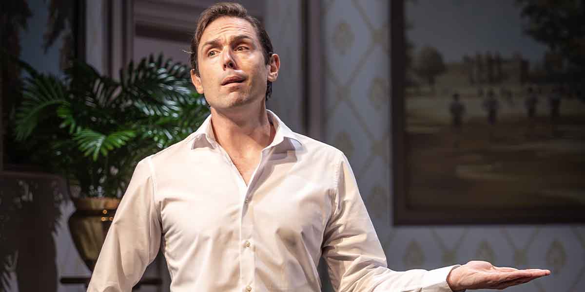 A man in a white shirt gestures expressively, with a green plant and artwork in the background.