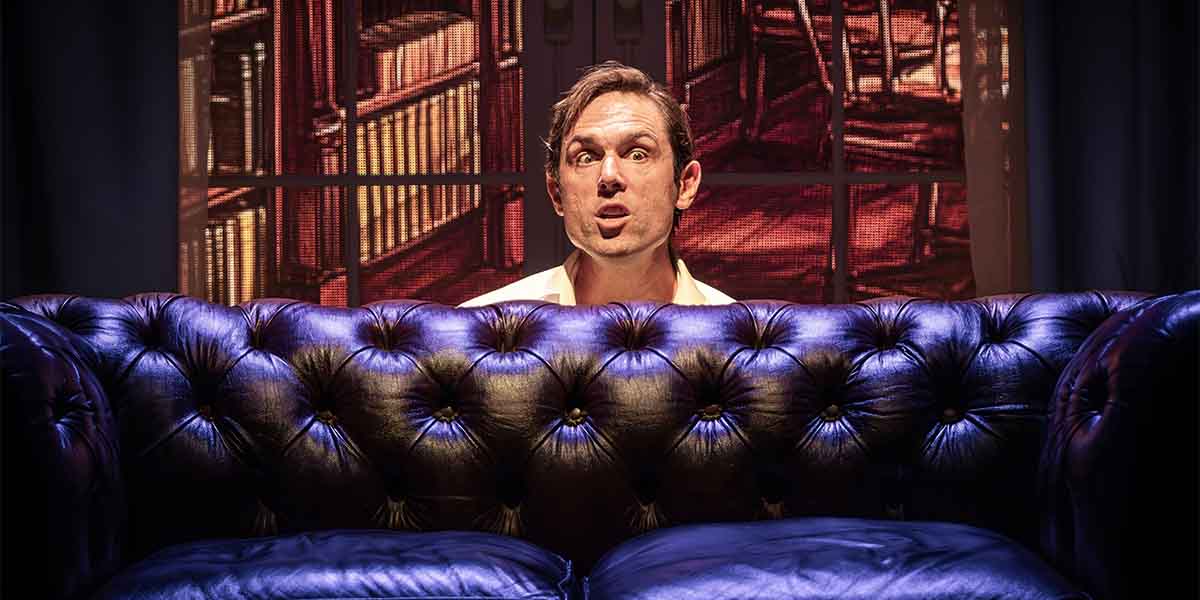 A man with short hair sits behind a dark blue tufted sofa, looking surprised against a vibrant background.
