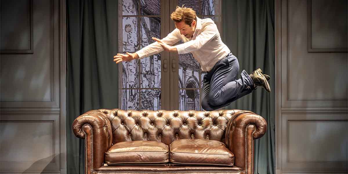 A man in a white shirt leaps over a brown leather sofa, with curtains and a window backdrop.