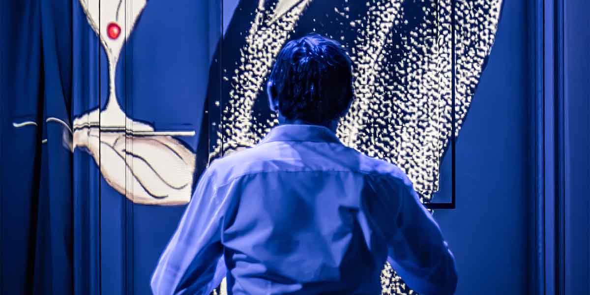 A person in a white shirt stands facing a large, illuminated abstract projection against a dark background.
