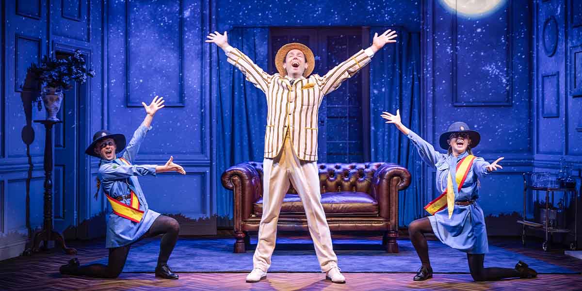 Three performers on stage: a man in striped suit and two women in blue outfits, all posing energetically.