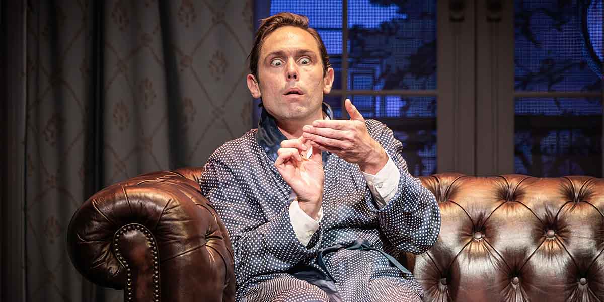 A man in a polka dot robe sits on a brown leather sofa, looking surprised and gesturing with his hands.