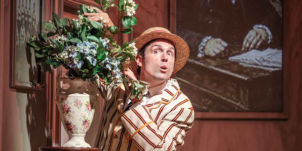 A man in a striped jacket and straw hat poses playfully beside a vase of flowers.