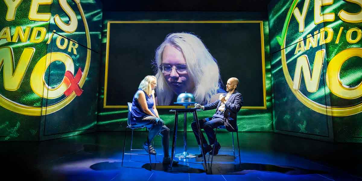 Derren Brown sits at a table with a woman, while a large screen displays her face.