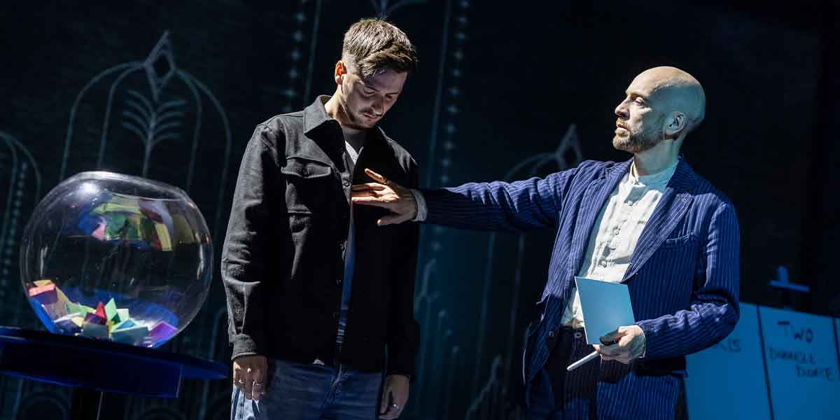 Derren Brown gestures towards a participant, who stands with a serious expression beside a glass bowl.