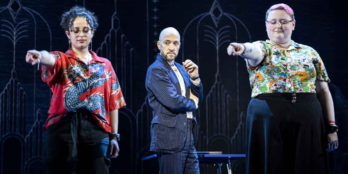 Derren Brown stands centre stage, flanked by two participants pointing, against a decorative backdrop.