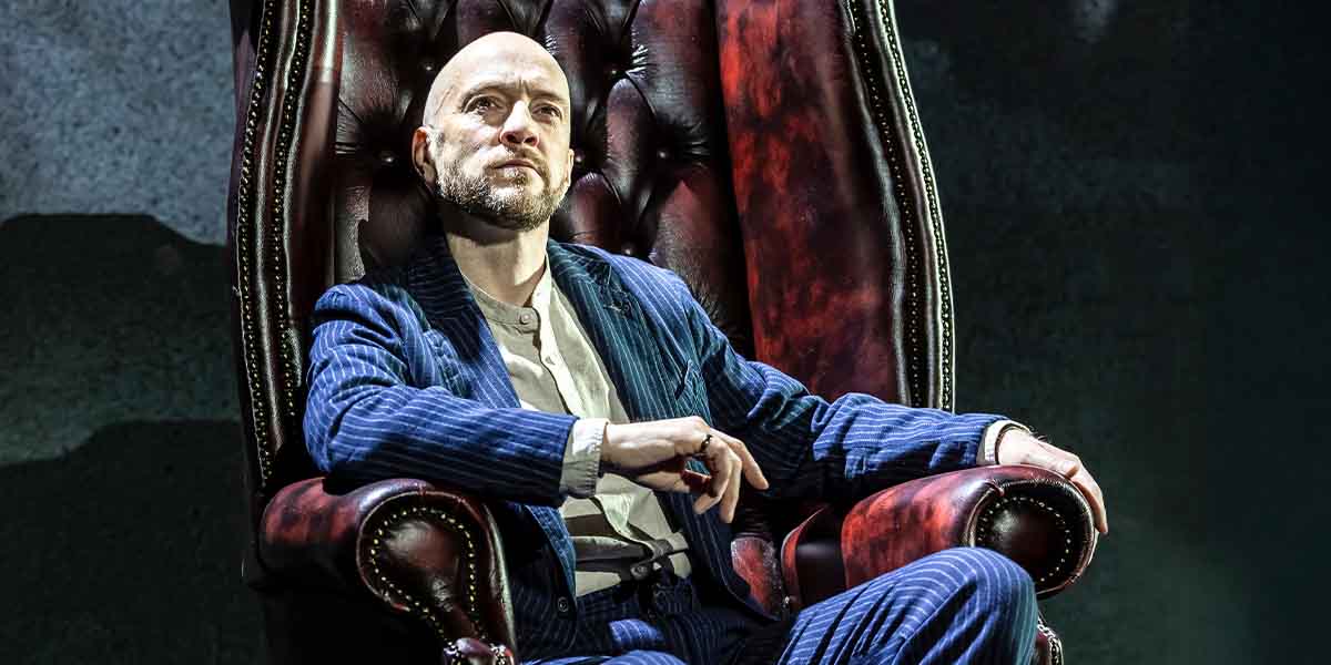 Derren Brown sits confidently in a large, ornate armchair, wearing a blue pinstripe suit and white shirt.
