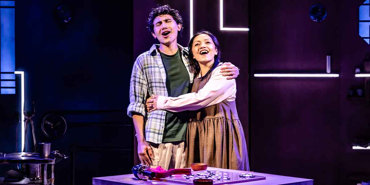 Two performers embrace and sing joyfully on stage, surrounded by a purple-lit backdrop and props.