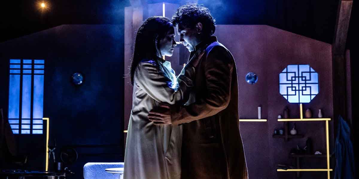A man and woman embrace closely, illuminated by blue lighting, in a stylised interior setting.