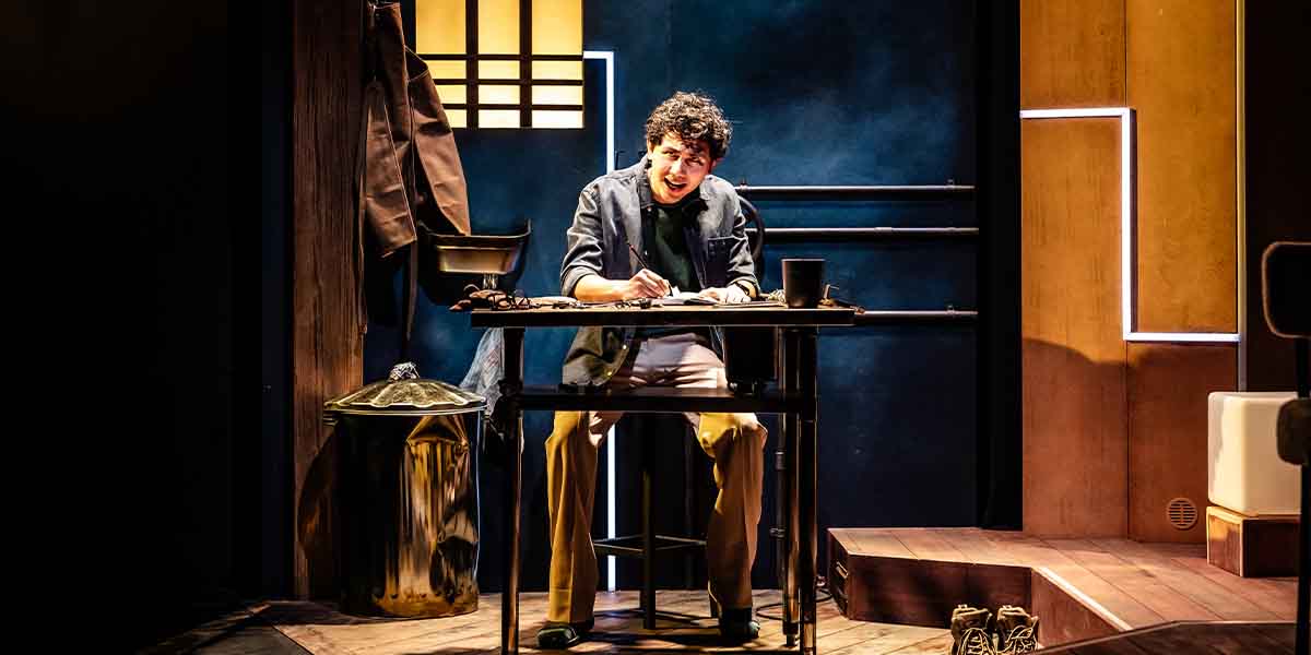 A young man sits at a desk, writing intently, with a trash can and coat visible nearby.