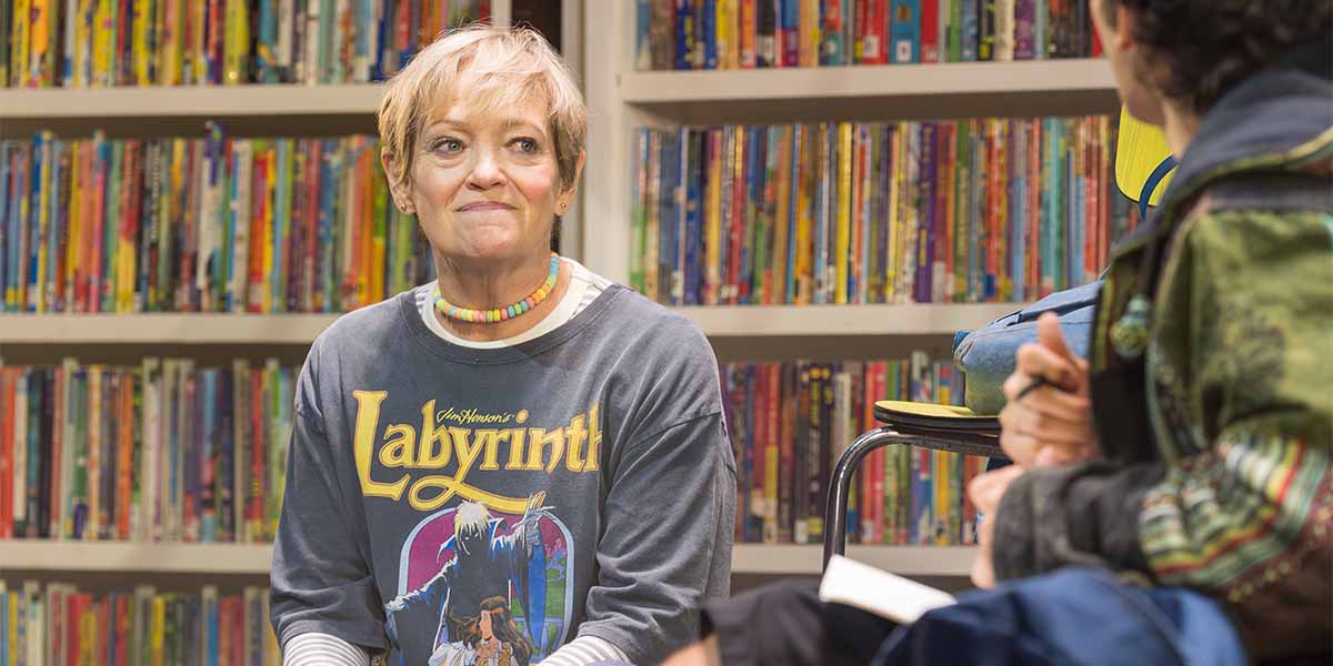 Maria Friedman as Kimberly Levaco, wearing a "Labyrinth" shirt, smiles in front of a colourful bookshelf.