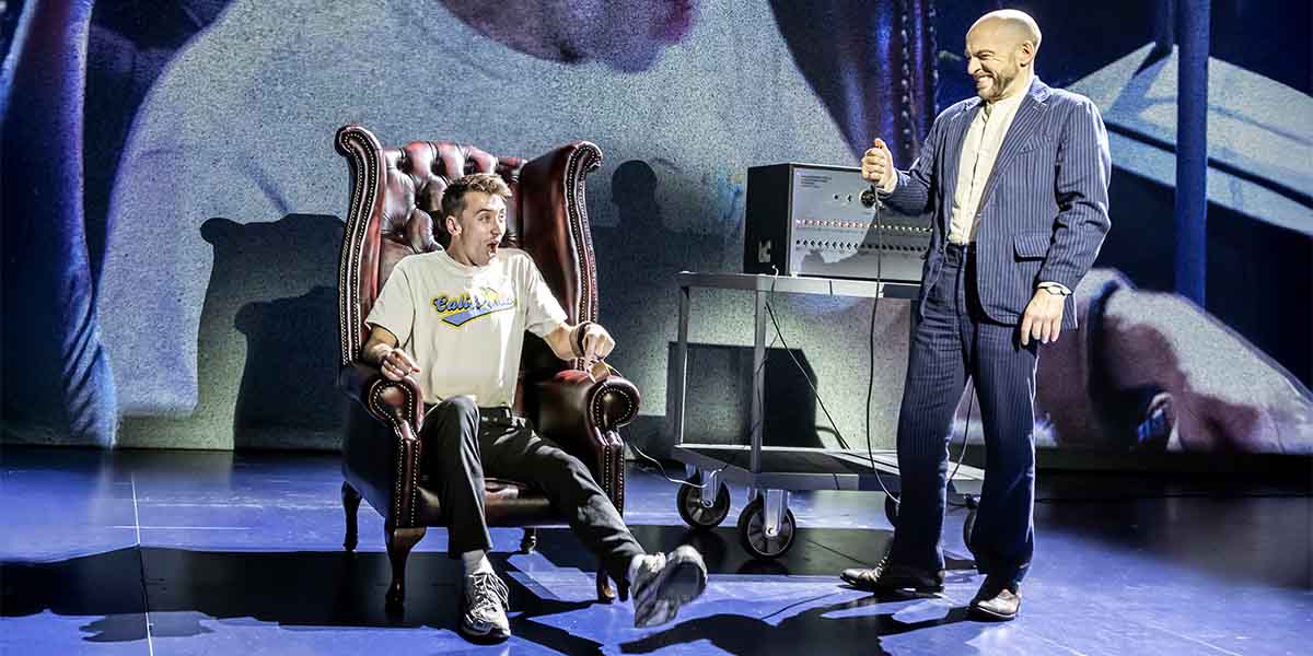 Derren Brown stands beside a surprised participant in a chair, with a control panel in the background.