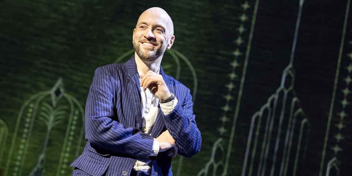 Derren Brown smiles confidently, wearing a striped suit, against a textured green background with geometric designs.