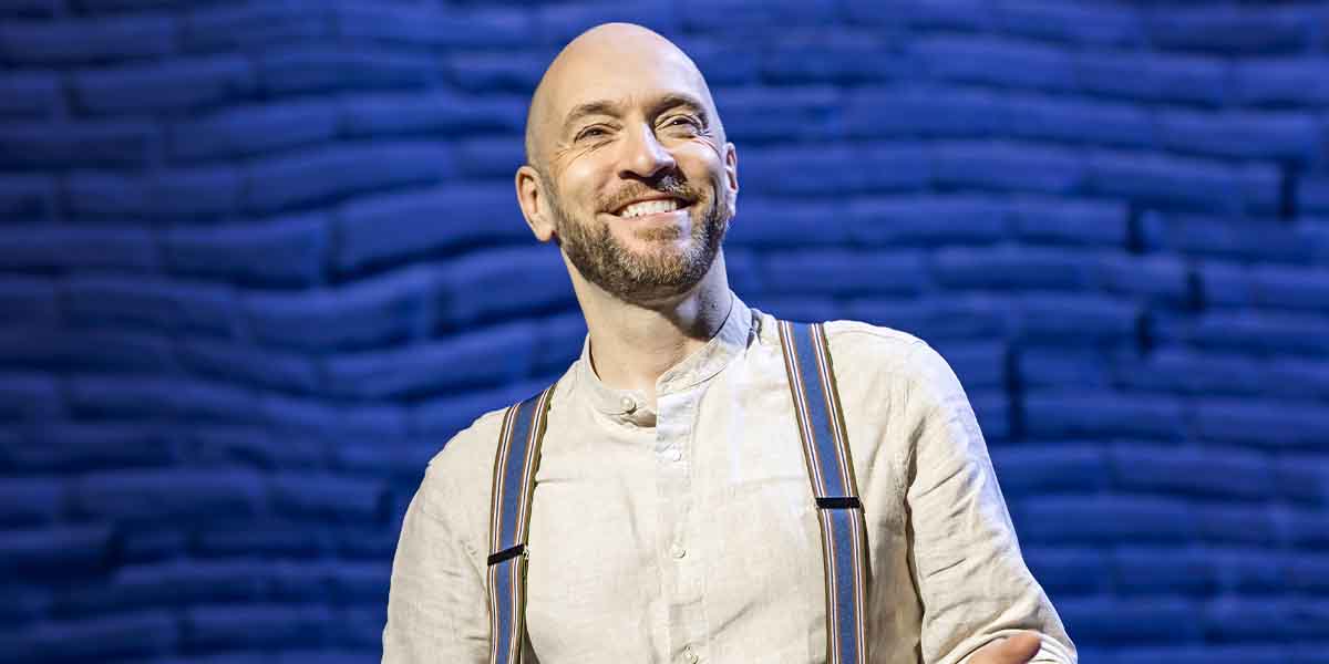 Derren Brown smiles while gesturing, wearing a light shirt and striped suspenders against a blue backdrop.