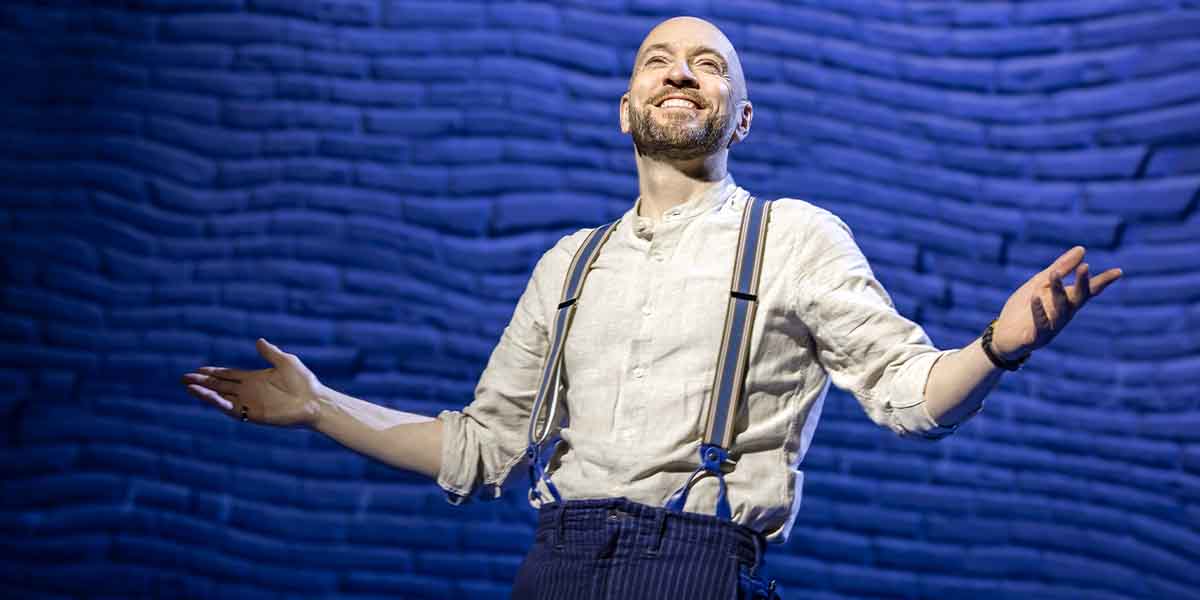 Derren Brown stands on stage, smiling with arms outstretched, against a textured blue background.