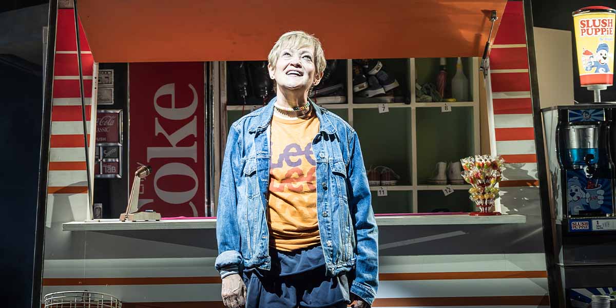 Maria Friedman as Kimberly Levaco, wearing a vibrant dress, stands confidently on stage with dramatic lighting.