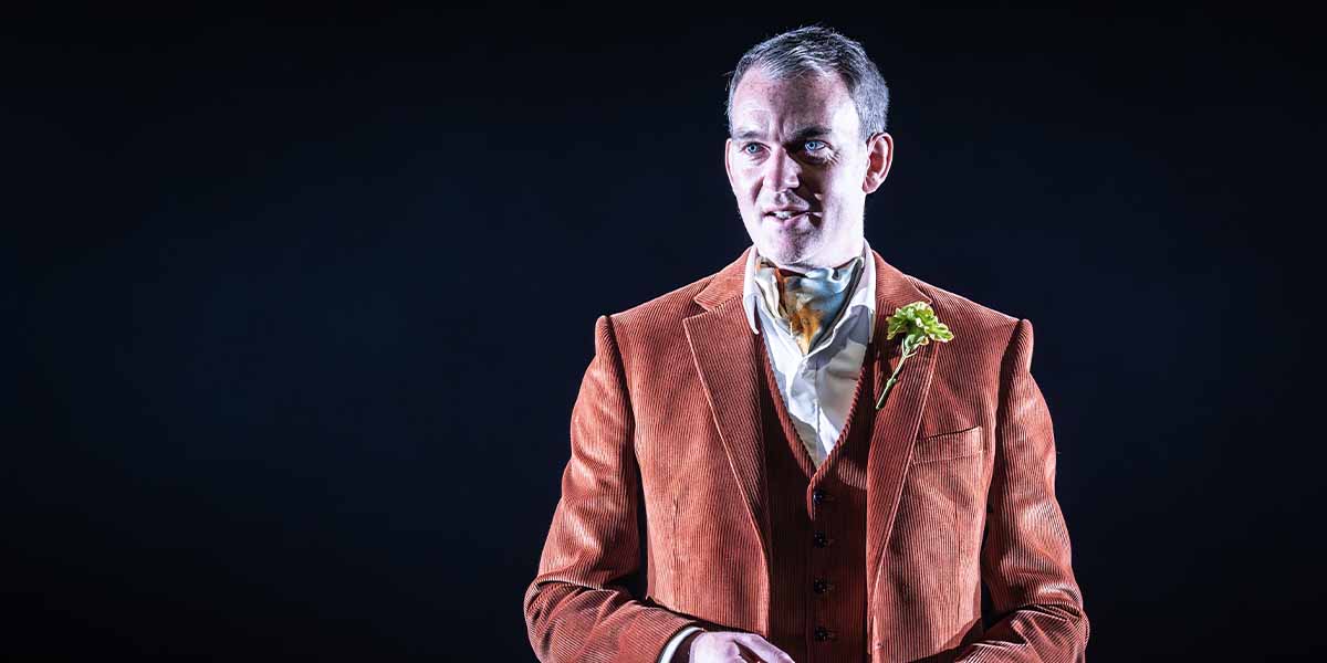 Alastair Whatley, wearing an orange corduroy suit with a green flower, speaks against a dark background.