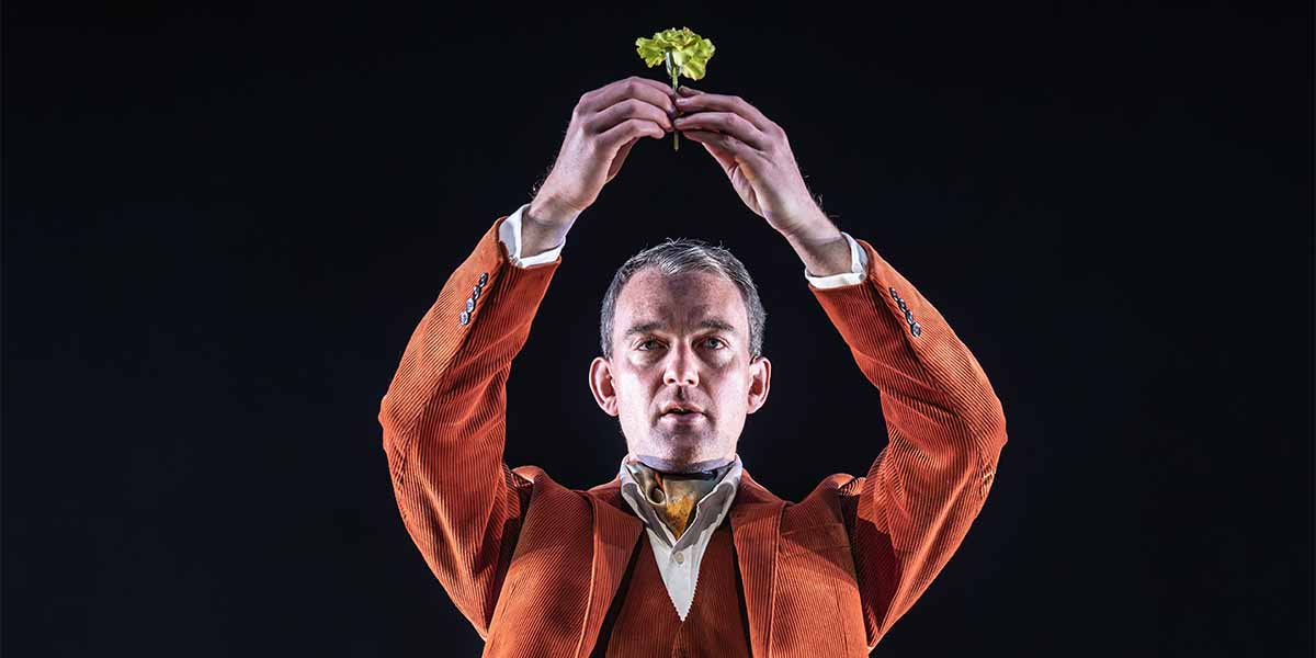 Alastair Whatley holds a green flower above his head, wearing an orange suit against a dark background.