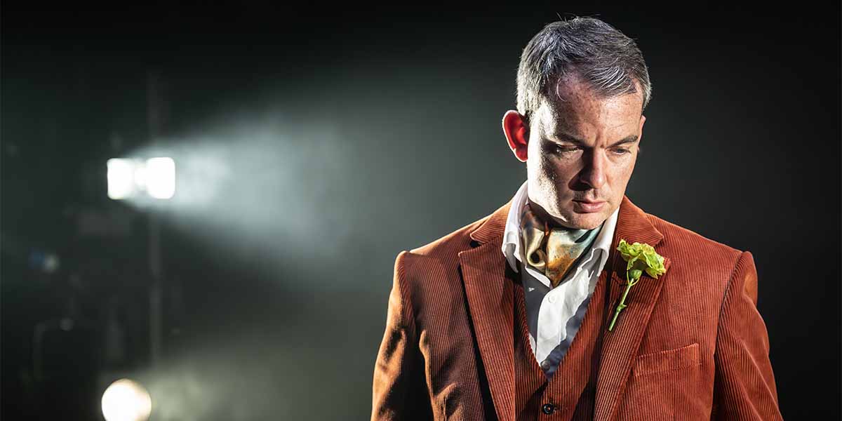 Alastair Whatley, dressed in a brown velvet suit with a green flower, gazes downward thoughtfully.