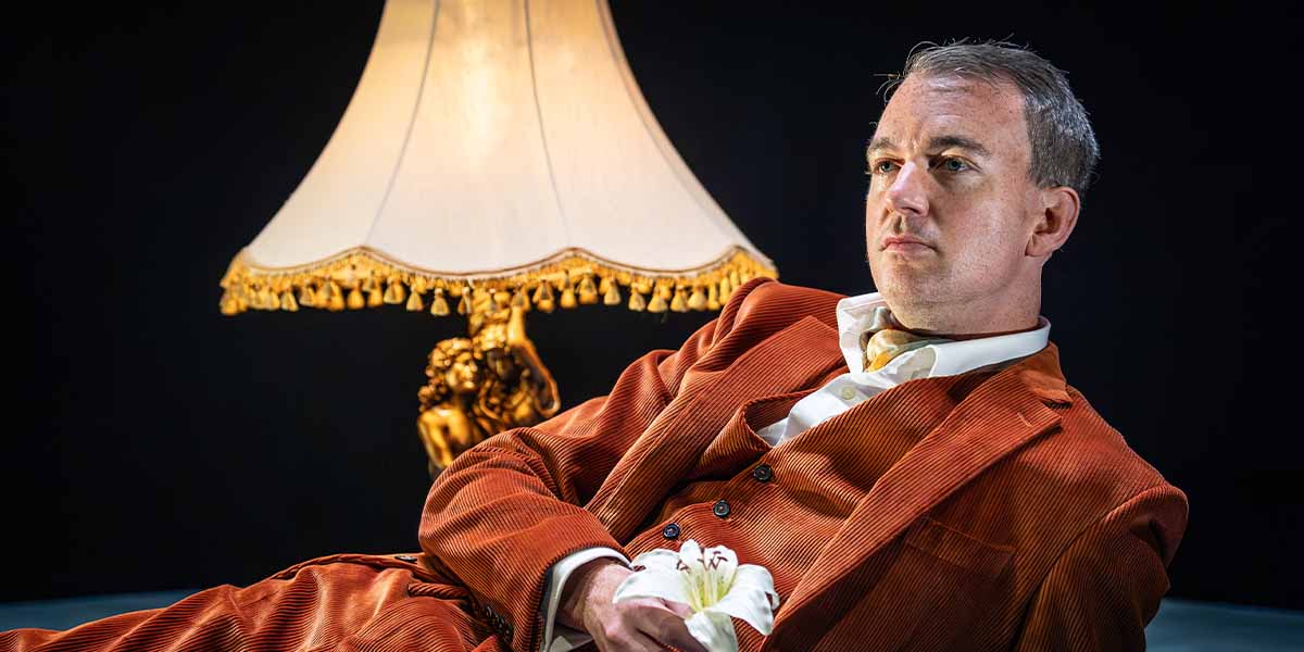 Alastair Whatley performs on stage, dressed in a formal suit, with dramatic lighting and a dark backdrop.