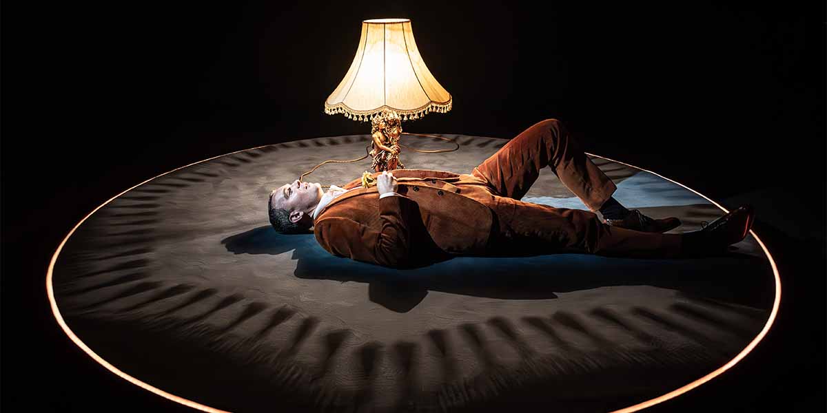 Alastair Whatley lies on a circular stage, illuminated by a lamp, wearing a brown suit.