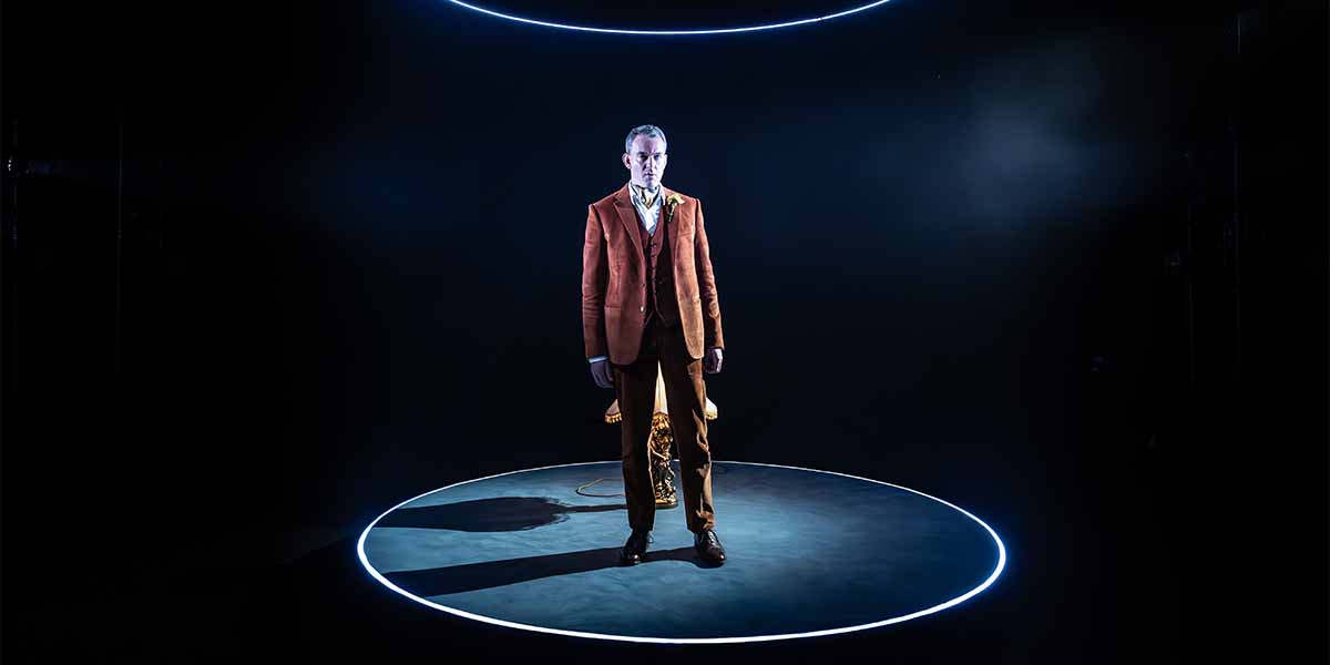 Alastair Whatley stands centre stage in a brown suit, illuminated by a circular light against a dark background.