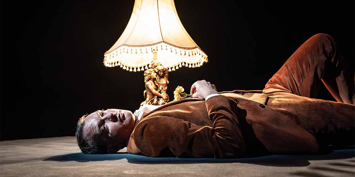 Alastair Whatley lies on the floor, illuminated by a lamp, wearing a brown corduroy suit.