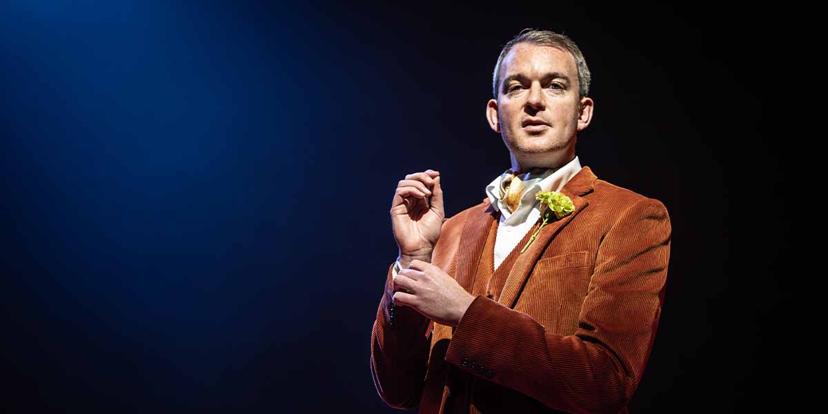 Alastair Whatley, wearing a brown corduroy jacket, gestures thoughtfully against a dark blue background.