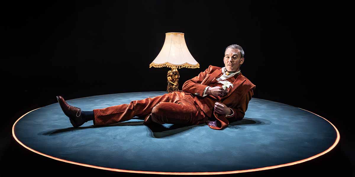 Alastair Whatley lounges on a circular stage, wearing an orange suit, beside a lamp.