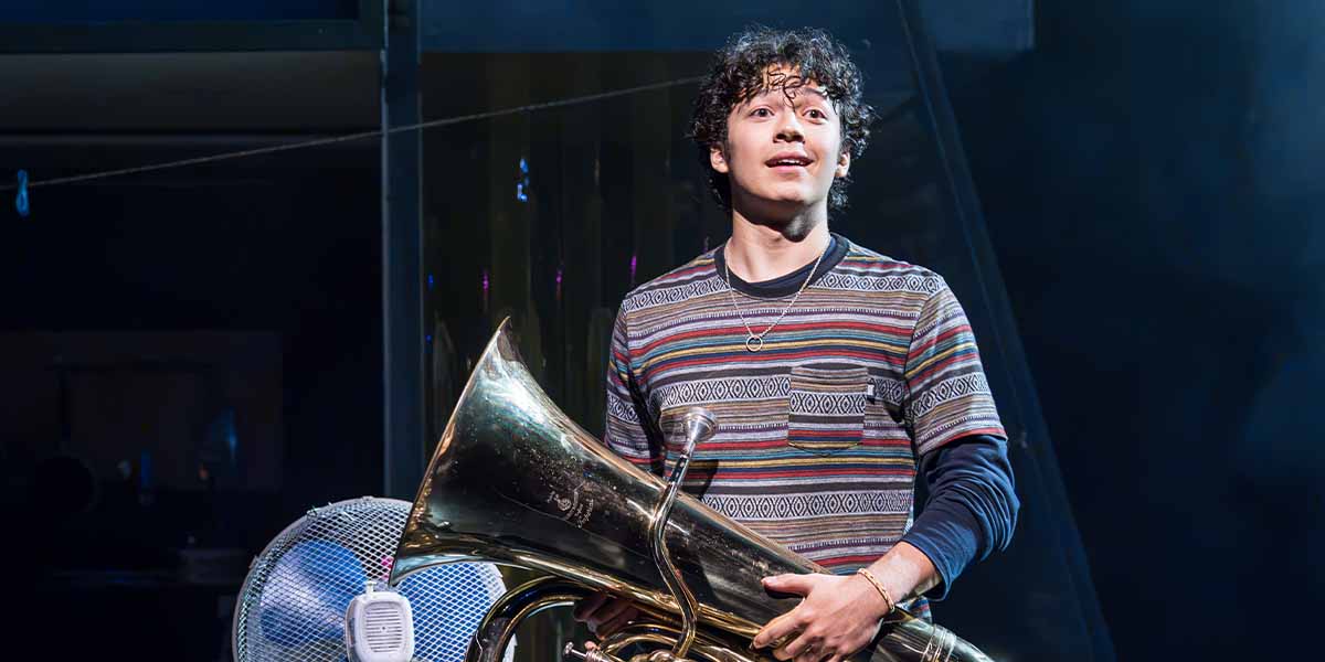 Gilli Jones as Seth Weetis holds a tuba, smiling, wearing a striped shirt against a dark background.
