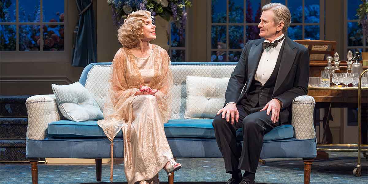 Christine Baranski as Judith Bliss and Charles Edwards as Richard Greatham sit on a blue sofa, smiling.
