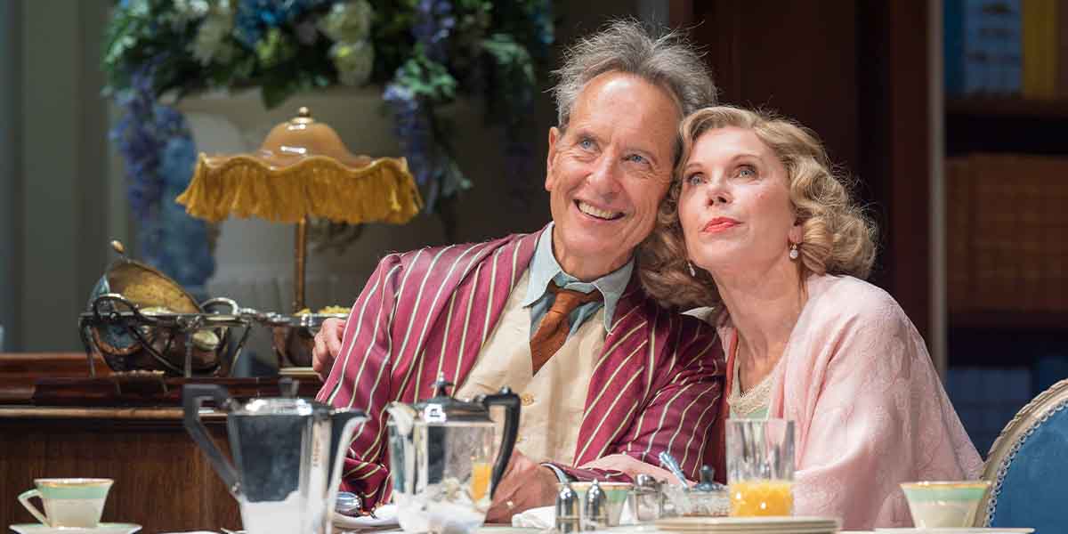 Richard E. Grant and Christine Baranski smile together at a table, surrounded by elegant tableware and decor.