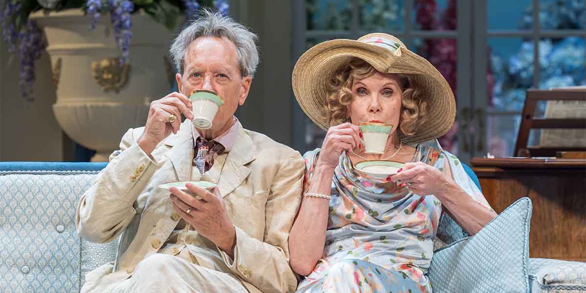 Richard E. Grant and Christine Baranski sit on a sofa, sipping tea, dressed in elegant attire.