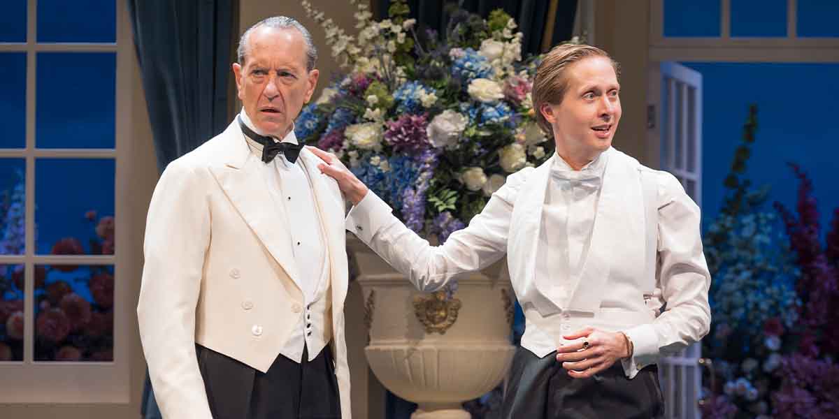 Richard E. Grant and Will Close, dressed in formal attire, interact in front of a floral backdrop.