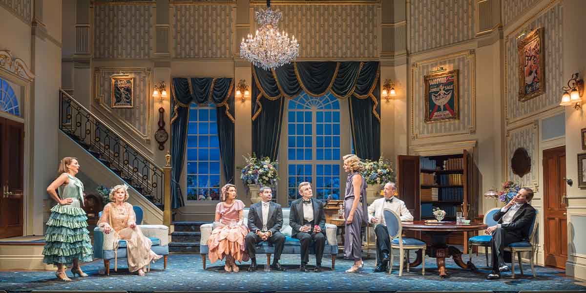 A theatrical scene featuring six characters in elegant attire, set in a lavishly decorated room.