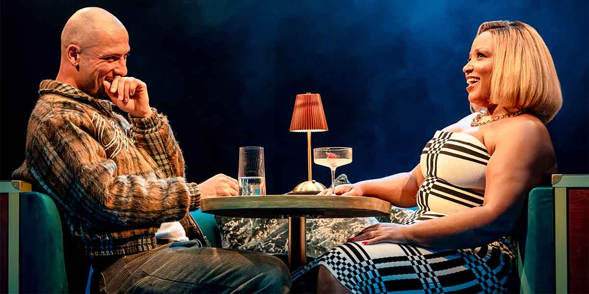 Jamael Westman and Marisha Wallace sit at a table, smiling, with drinks and a lamp between them.