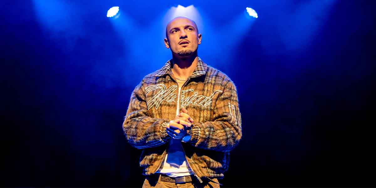 Jamael Westman as Dallas, standing with clasped hands, wearing a patterned jacket against a blue-lit background.