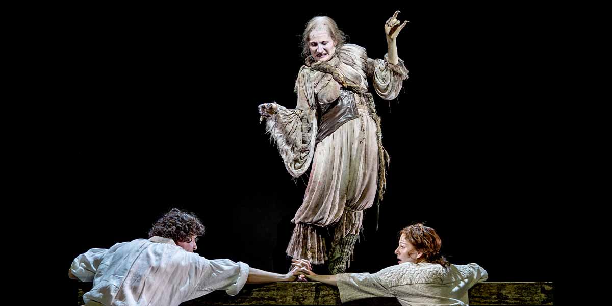 Kate Fleetwood, in tattered costume, gestures dramatically while Laura Pitt-Pulford and John Dagleish reach towards her.