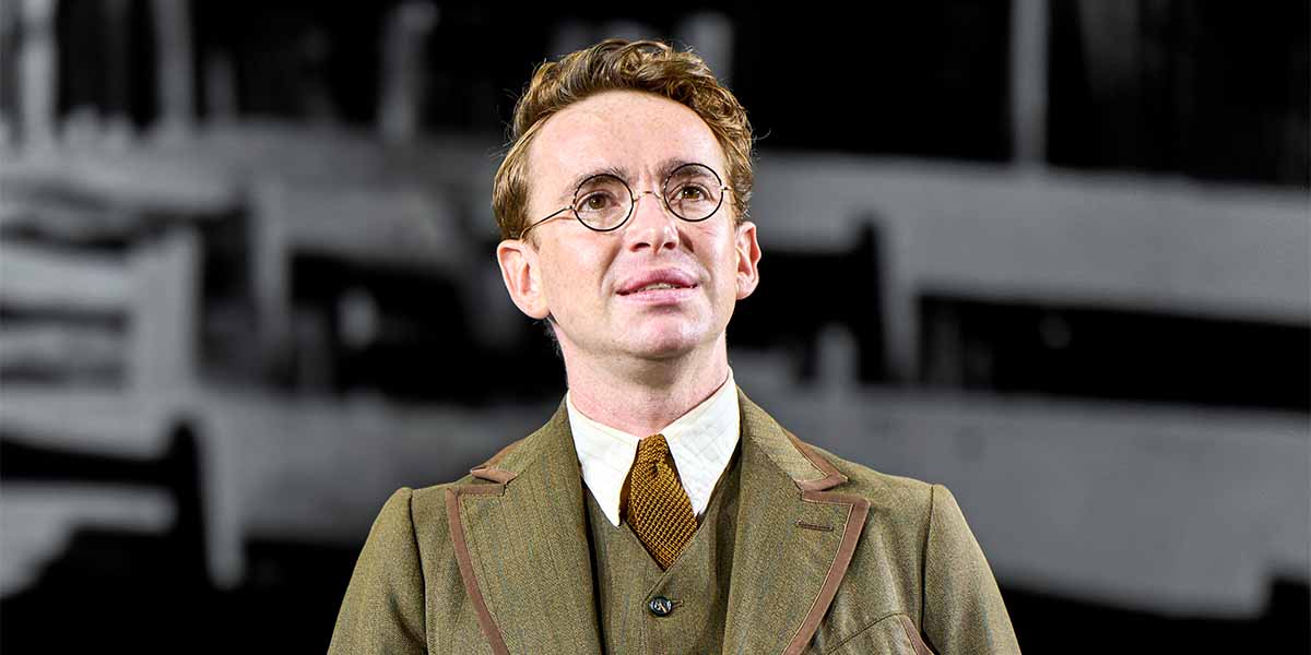 Laurie Kynaston, wearing a brown suit and glasses, gazes thoughtfully against a blurred background.