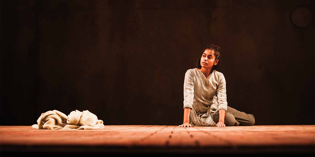 Linnea Berthelsen, in a light grey outfit, sits on a wooden stage, gazing thoughtfully.