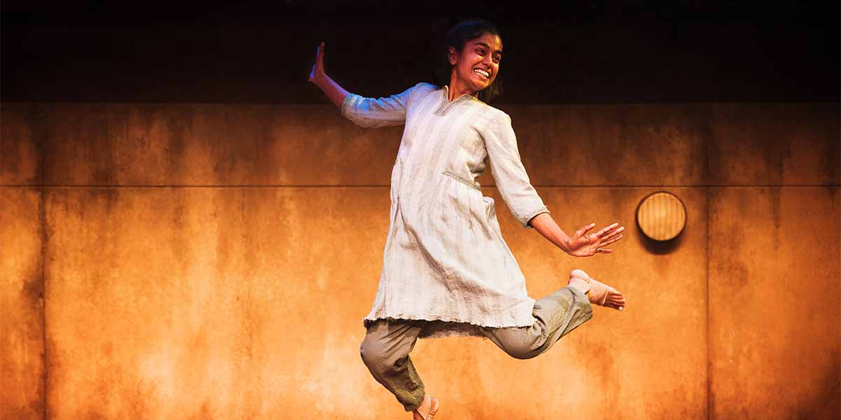 Linnea Berthelsen, in a light-coloured outfit, joyfully leaps on stage against a warm, textured backdrop.