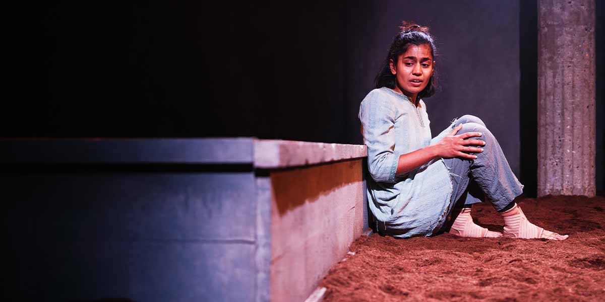 Linnea Berthelsen, in a light blue outfit, sits on sandy ground, looking distressed in a dimly lit scene.