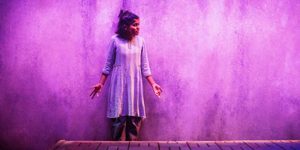 Linnea Berthelsen, in a light blue dress, gestures expressively against a purple wall backdrop.