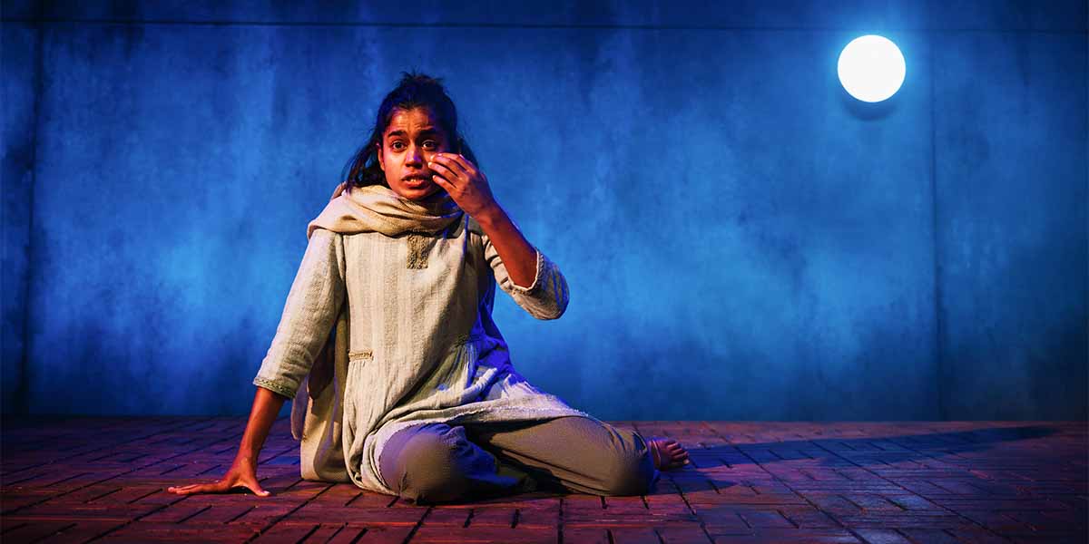 Linnea Berthelsen, in a light-coloured outfit, sits on a wooden stage, expressing emotion against a blue backdrop.