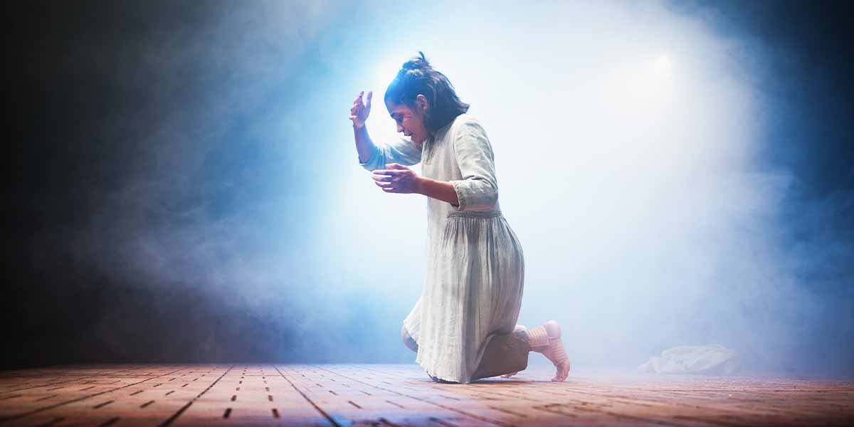 Linnea Berthelsen kneels on stage, wearing a light dress, surrounded by mist and dramatic lighting.