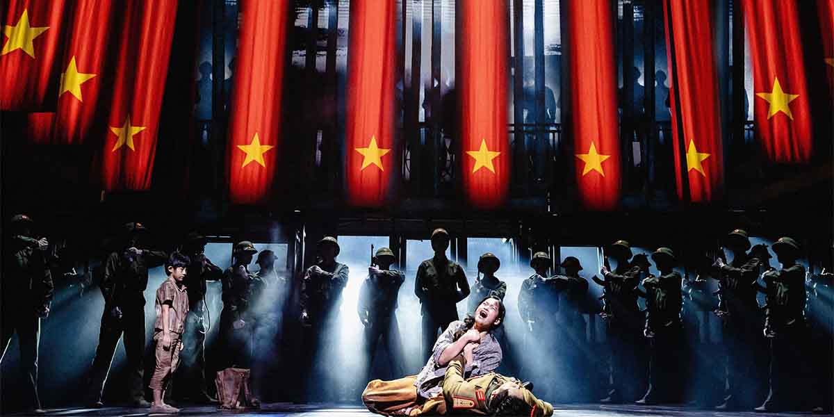 A child stands in front of a distressed performer, with red flags and soldiers in the background.